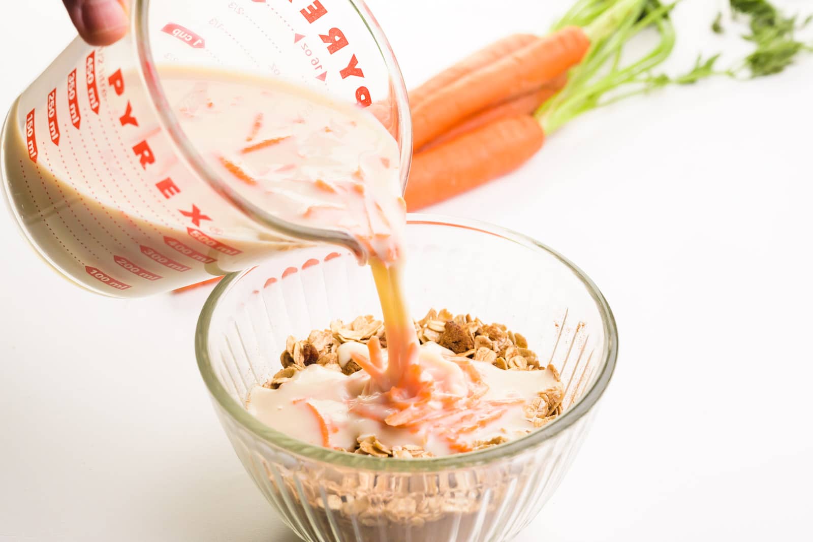 A hand holds a measuring cup pouring wet ingredients into a bowl with oats and spices. There are raw carrots in the background.