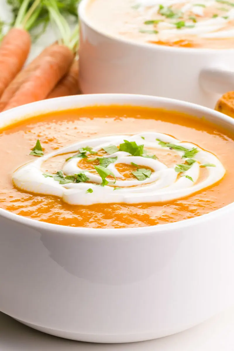 A bowl of carrot soup has cream drizzled across the top. There's another bowl behind it and some carrots on the side.