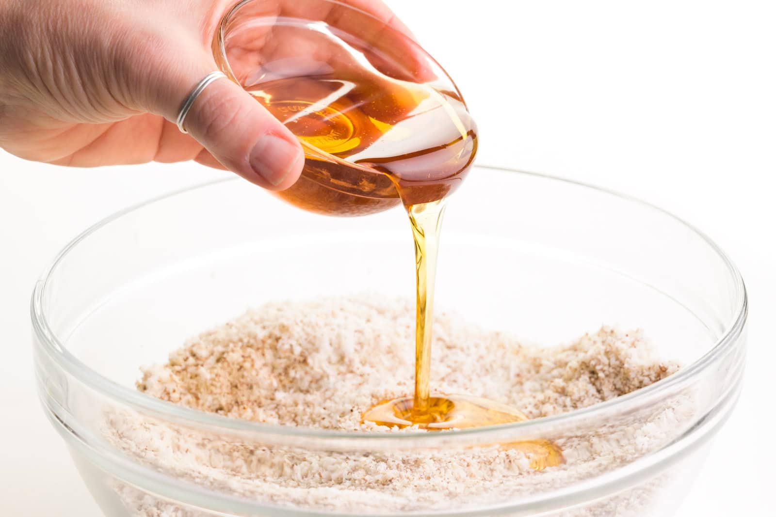 A hand holds a small bowl of syrup, pouring it into another bowl full of a coconut mixture.