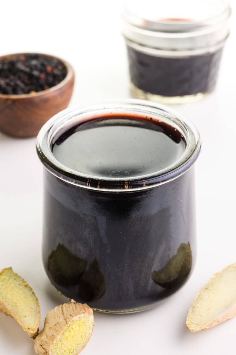 A small glass jar holds elderberry syrup. There are slices of fresh ginger in front of it. Behind it is a bowl of dried elderberries and more syrup.