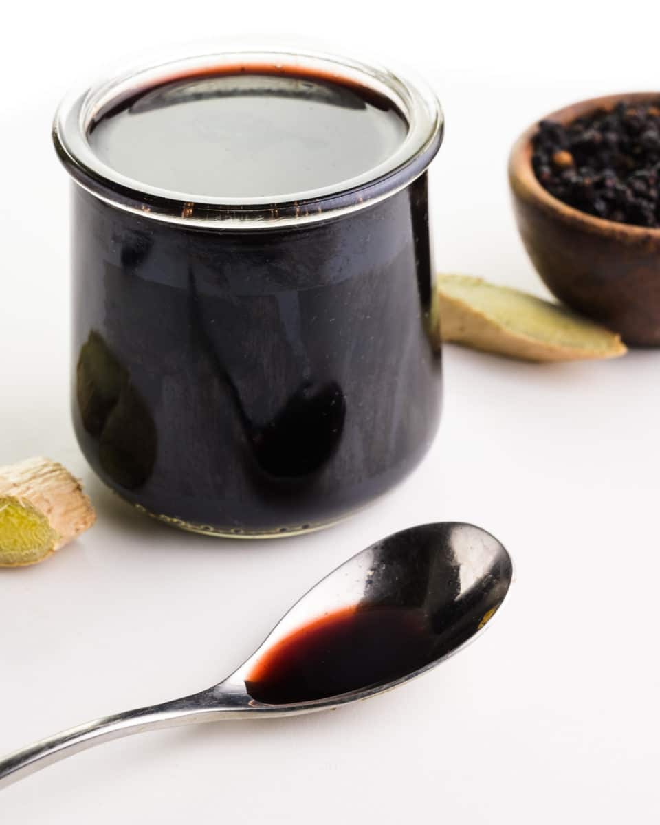 A spoon with elderberry syrup sits in front of a jar with more syrup. There are slices of fresh ginger and a bowl of dried elderberries in the background.