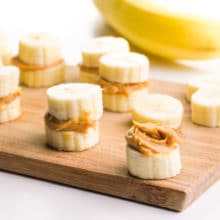 A cutting board holds several banana "sandwiches", with banana slices topped with peanut butter. There's a banana in the background.