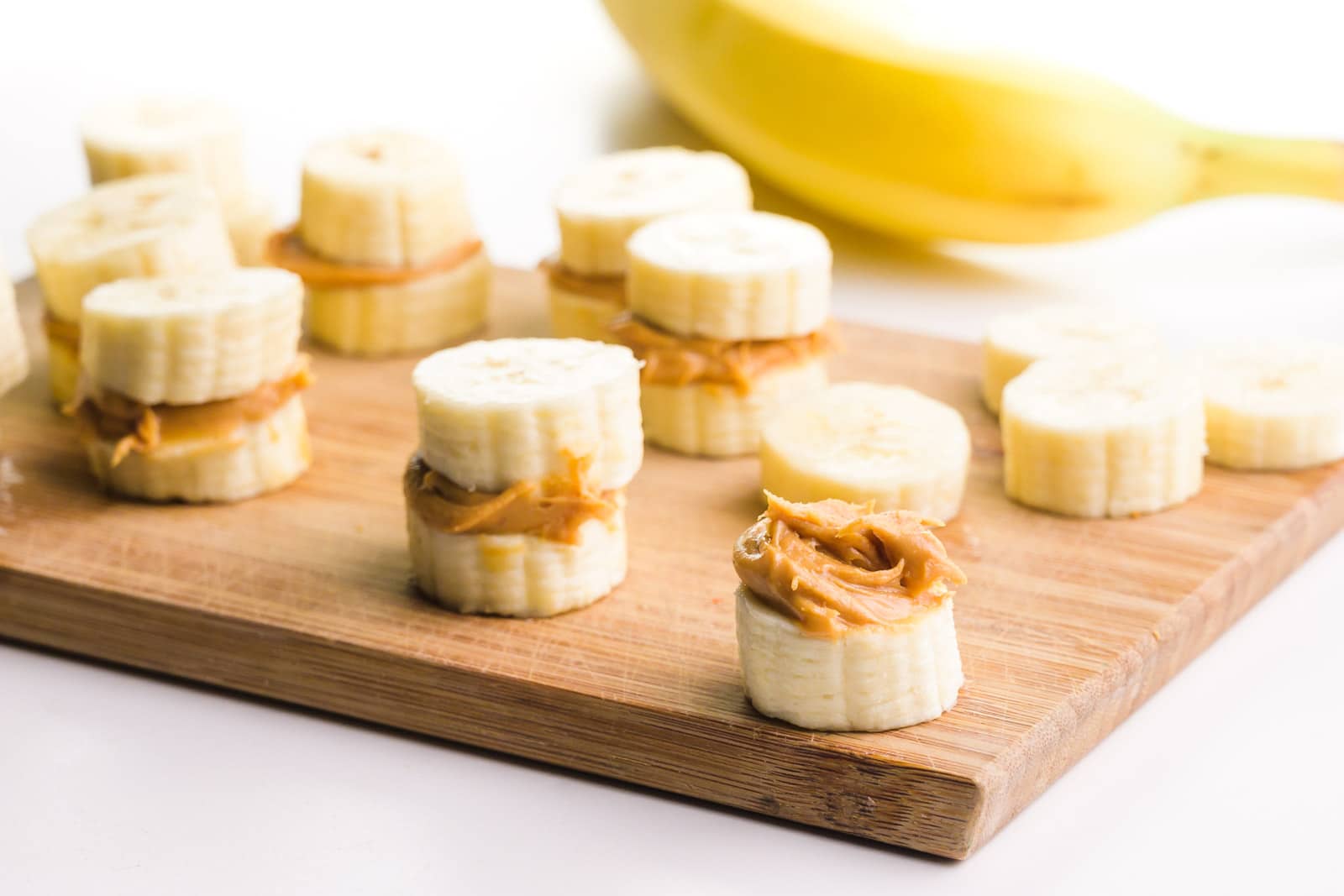 A cutting board holds several banana "sandwiches", with banana slices topped with peanut butter. There's a banana in the background.