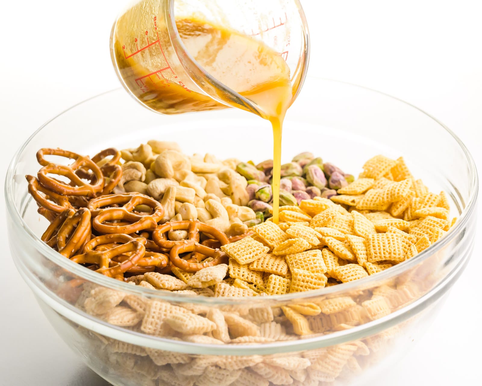 A butter mixture is being poured over snack mix cereal, pretzels, and nuts in a large bowl.