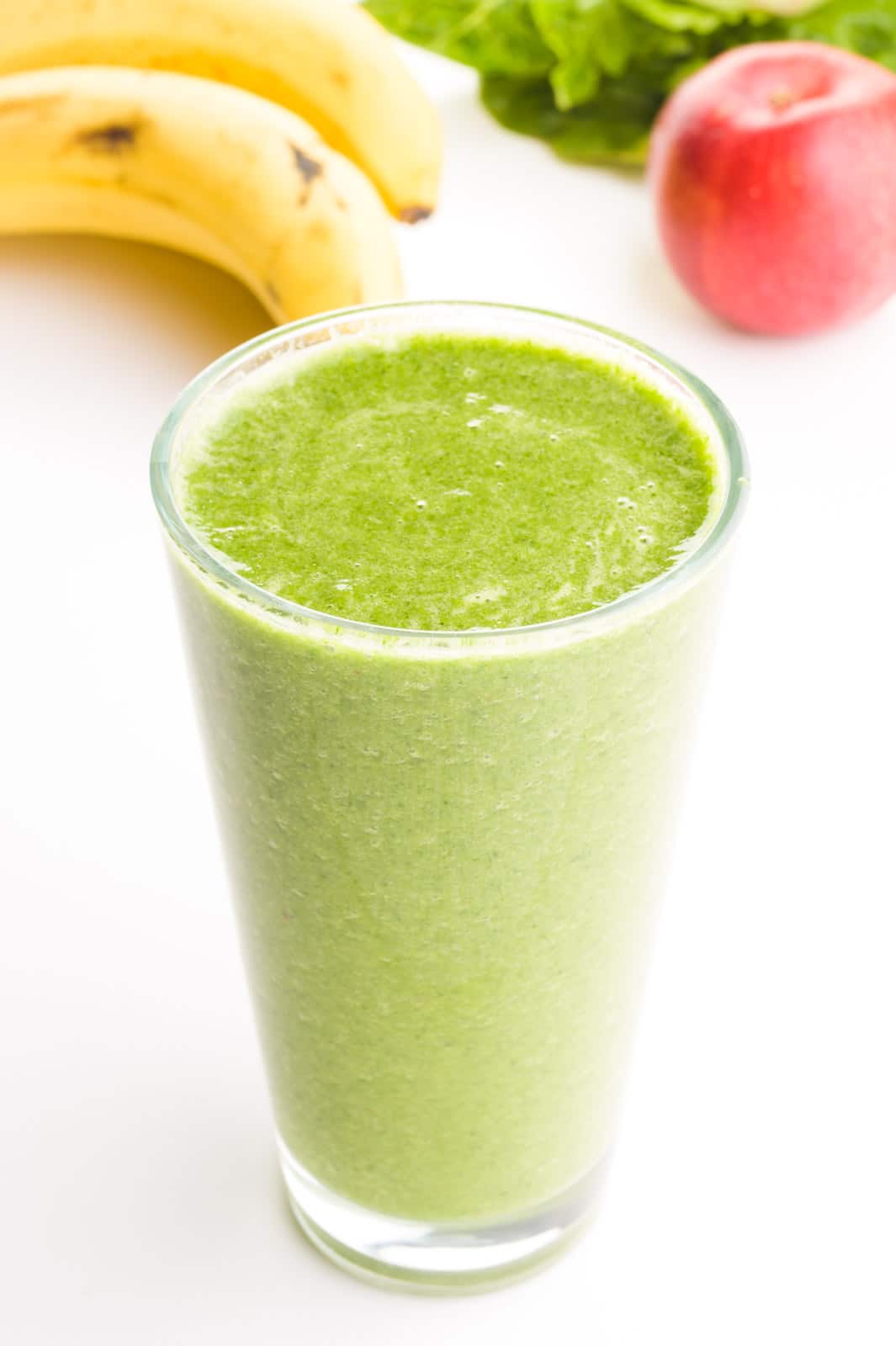 A glass if full of green smoothie. There's bananas, an apple, and greens in the background.