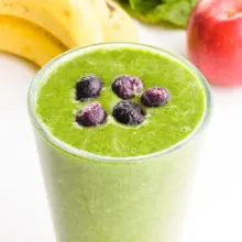 A glass holds bright green smoothie and blueberries on top. There are bananas with an apple and greens in the background.
