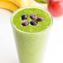 A green smoothie has blueberries on top. There are bananas, apples, and greens behind it.
