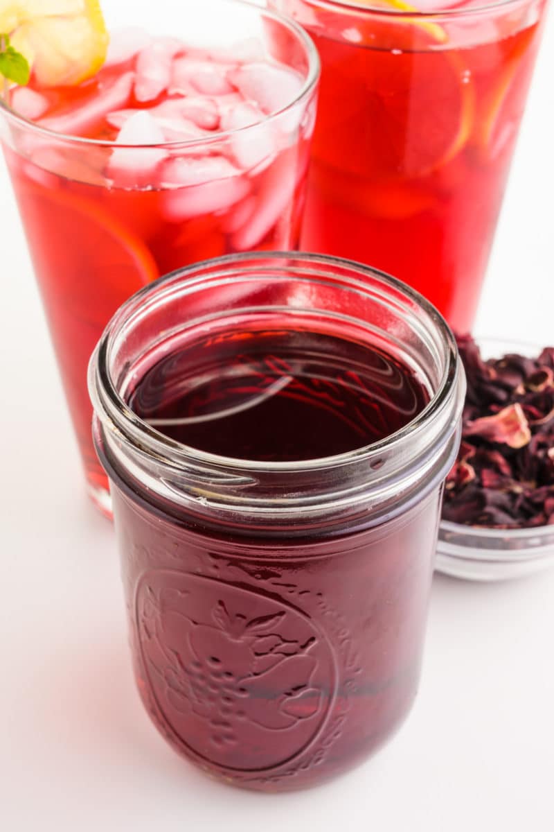 A mason jar holds hibiscus syrup with two glasses of the iced beverage behind it.