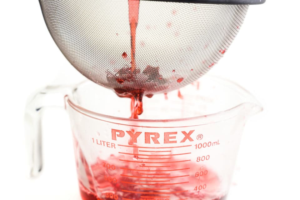 A pink liquid is being poured into a fine mesh strainer, with the liquid going into a glass pyrex measuring cup.