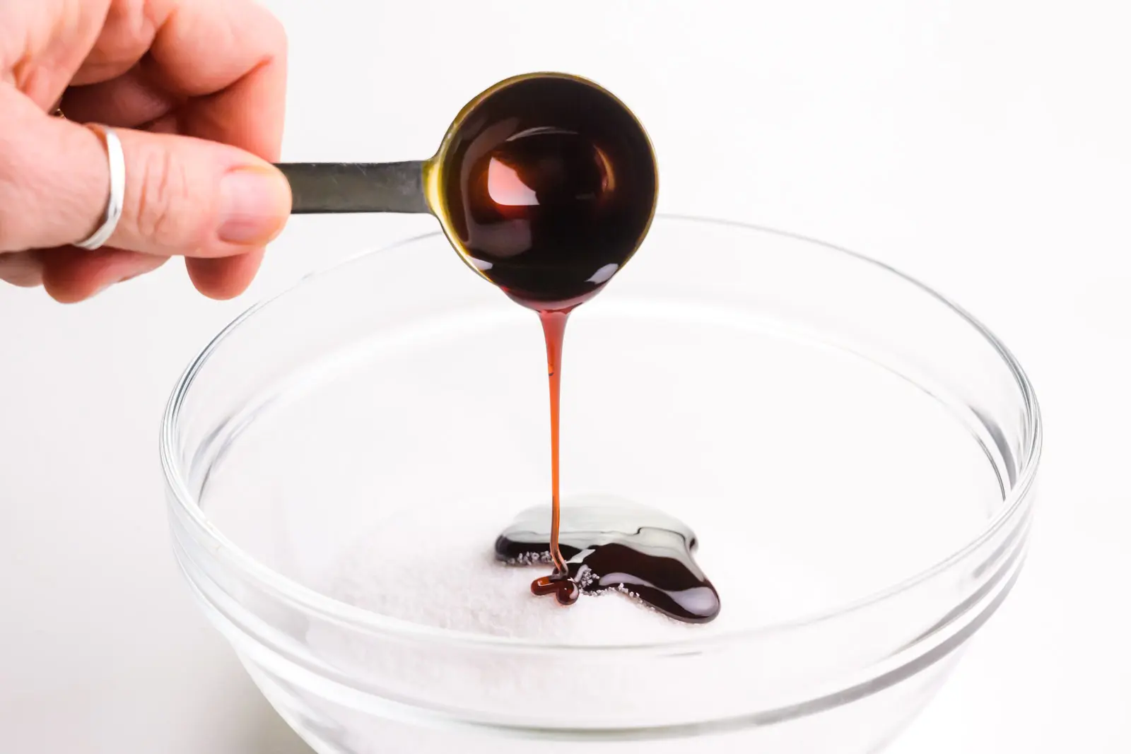 A hand holds a measuring spoon, drizzling molasses over a bowl of sugar.