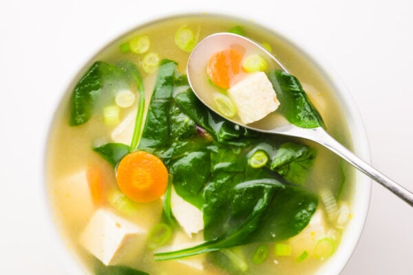 Looking down on a bowl of soup with a spoon in it. There are ingredients like greens, carrots, tofu, and green onions on top.
