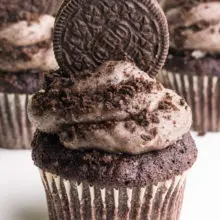 A cupcake has vegan Oreo frosting on top with an Oreo cookie on top. There are more cupcakes behind it.