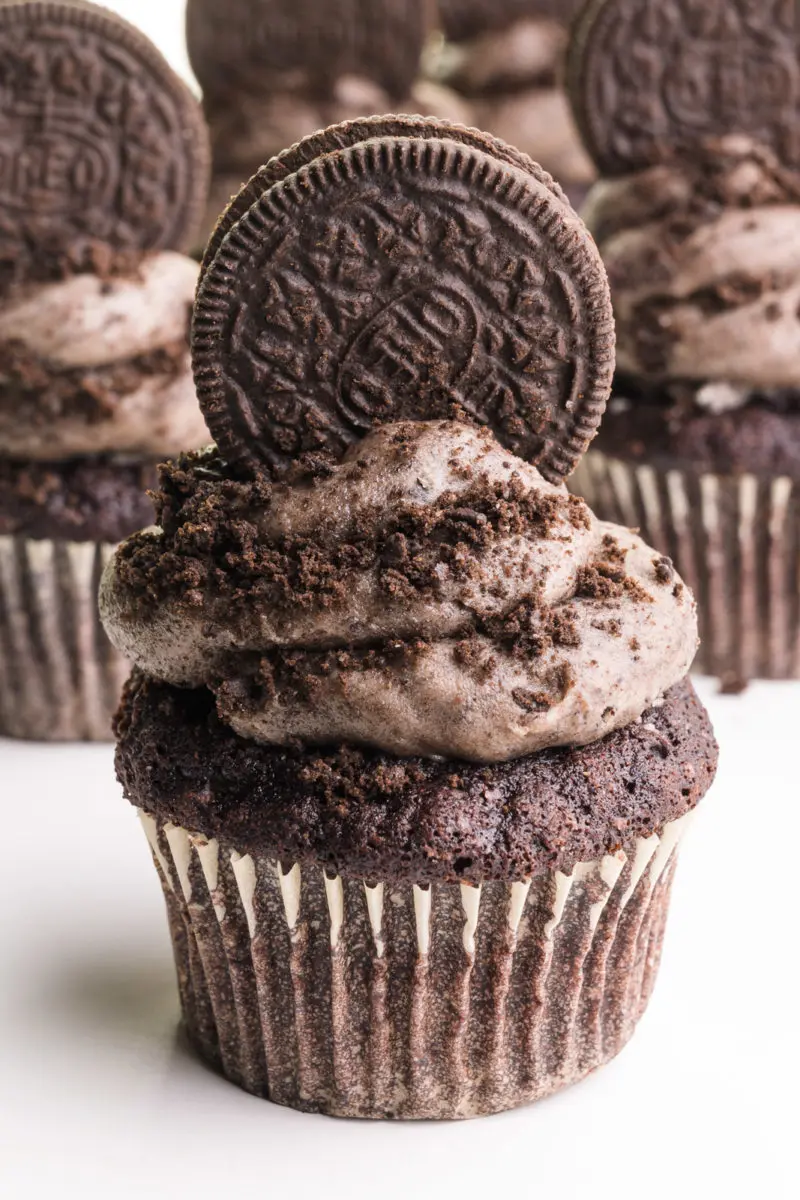 A cupcake has vegan Oreo frosting on top with an Oreo cookie on top. There are more cupcakes behind it.