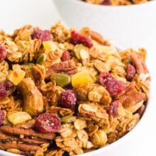 A bowl of peanut butter granola features dried cranberries, nuts, and more. There's another bowl with granola behind it.