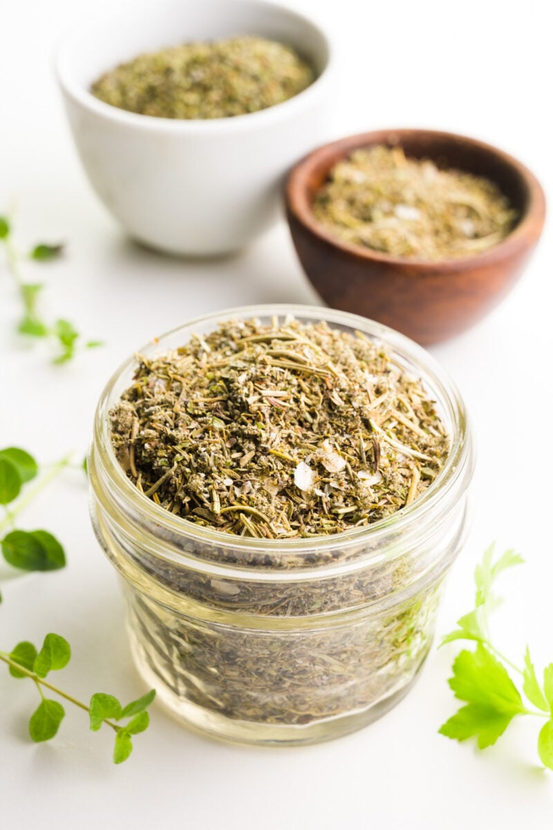 A jar of homemade poultry seasoning has fresh herbs around it and two bowls of more spices behind it.