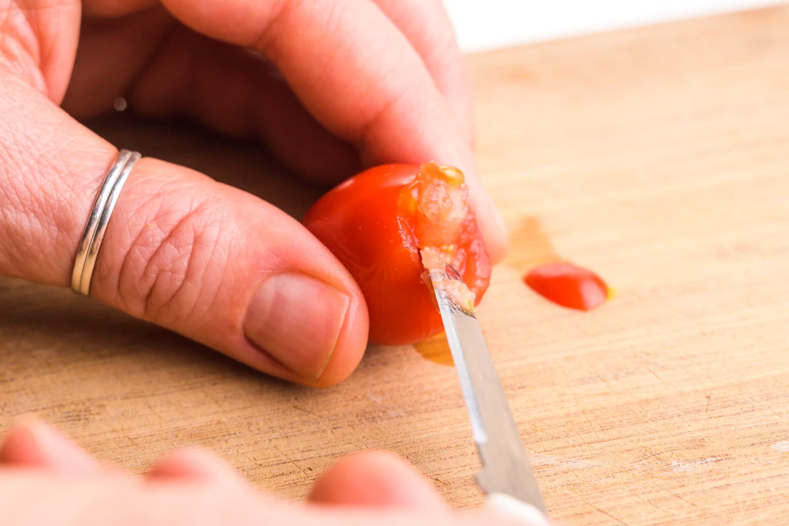 A hand holds a cherry tomato on its side on a cutting board. A paring knife is cutting out the insides of the tomato.