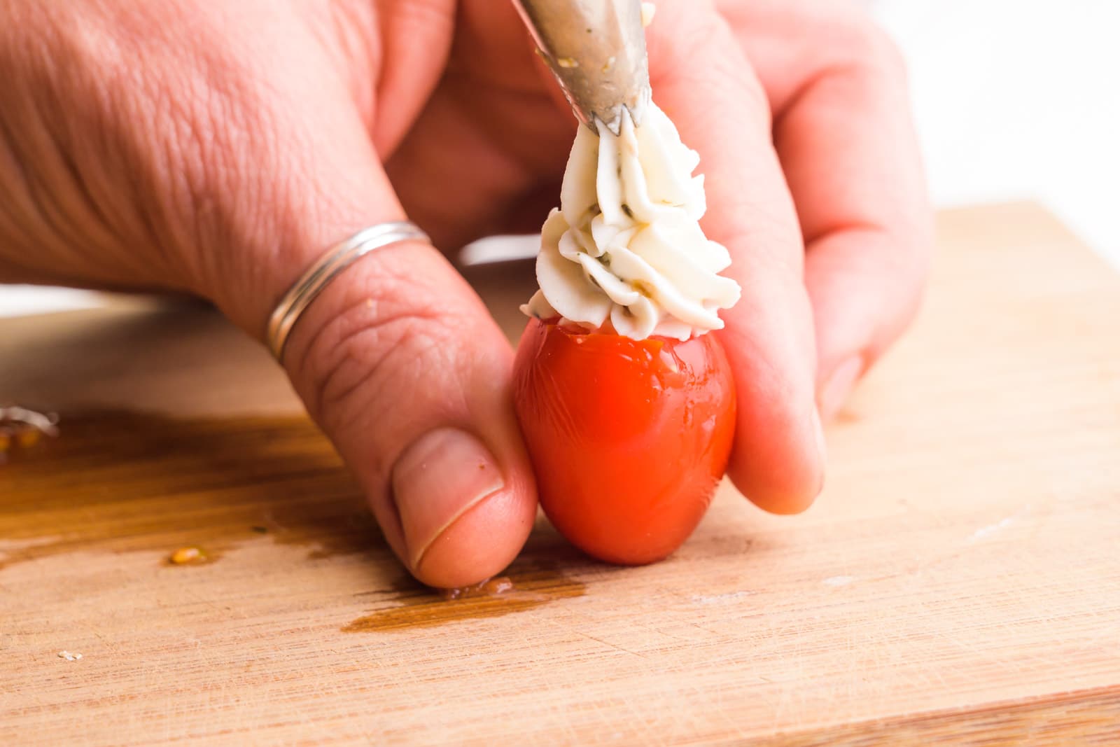 A hand holds a cherry tomato while using a piping bag to fill it with a cream cheese mixture.