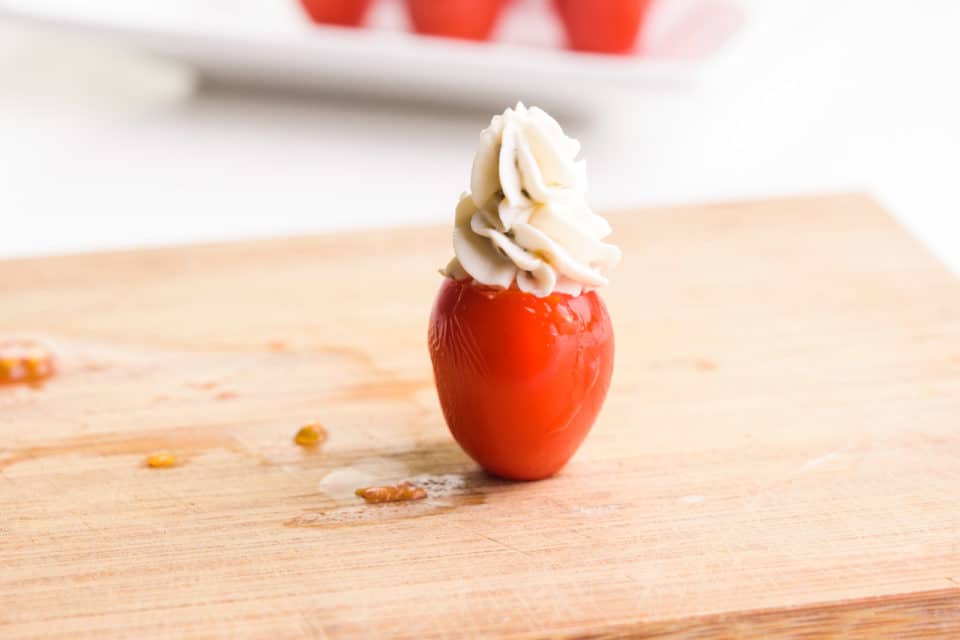 A single stuffed tomato stands upright on a cutting board, having just been filled with cream cheese. A tray of more tomatoes is in the background.