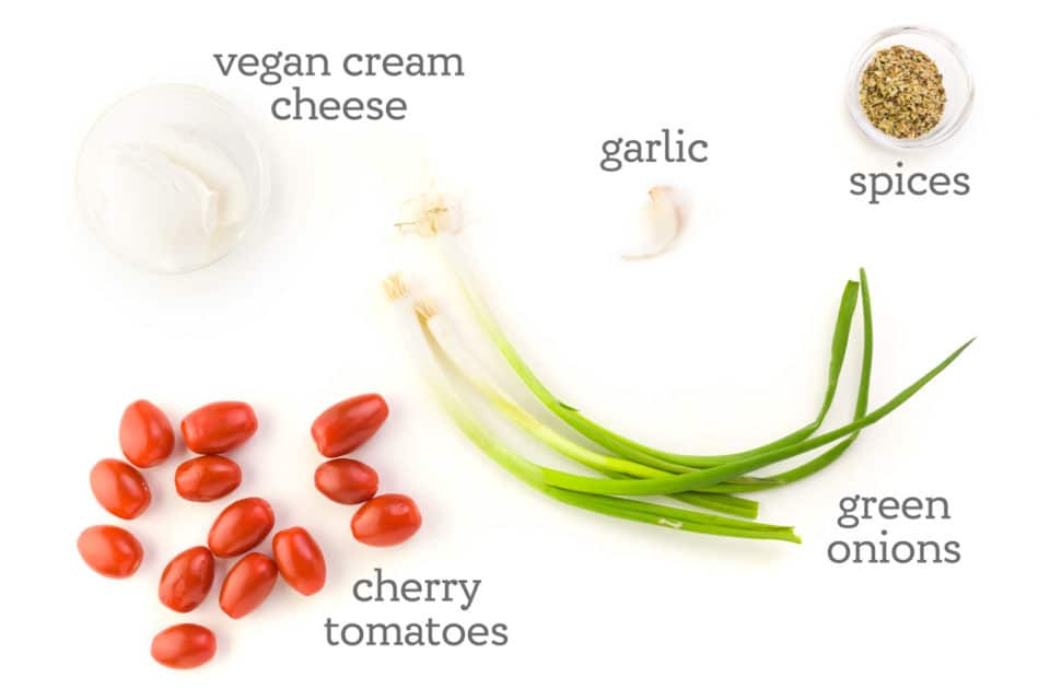 Ingredients are on a white counter. The labels around them read, "vegan cream cheese, garlic, spices, green onions, and cherry tomatoes."