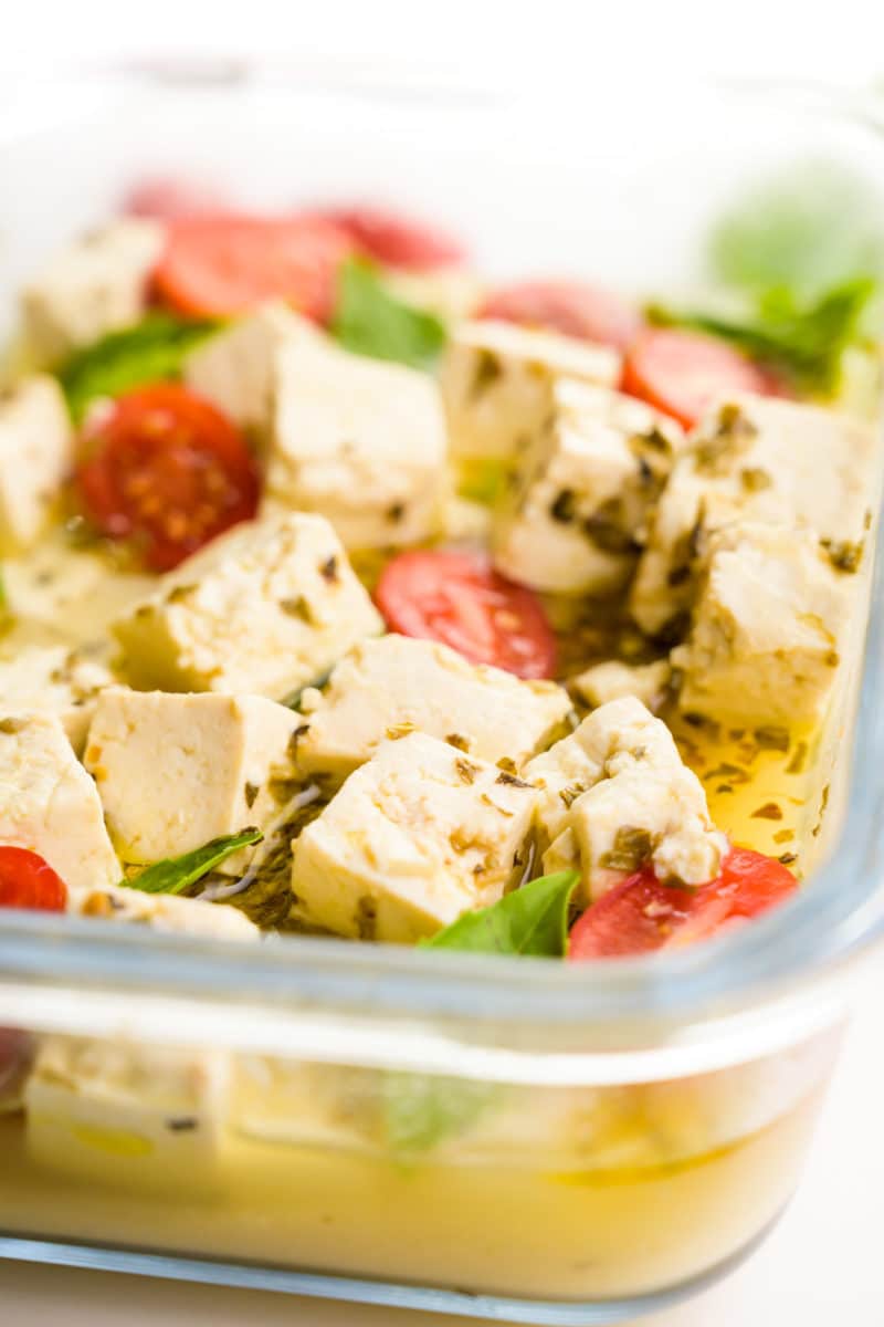 A glass container holds tofu feta with cherry tomatoes and basil leaves in a marinade. 