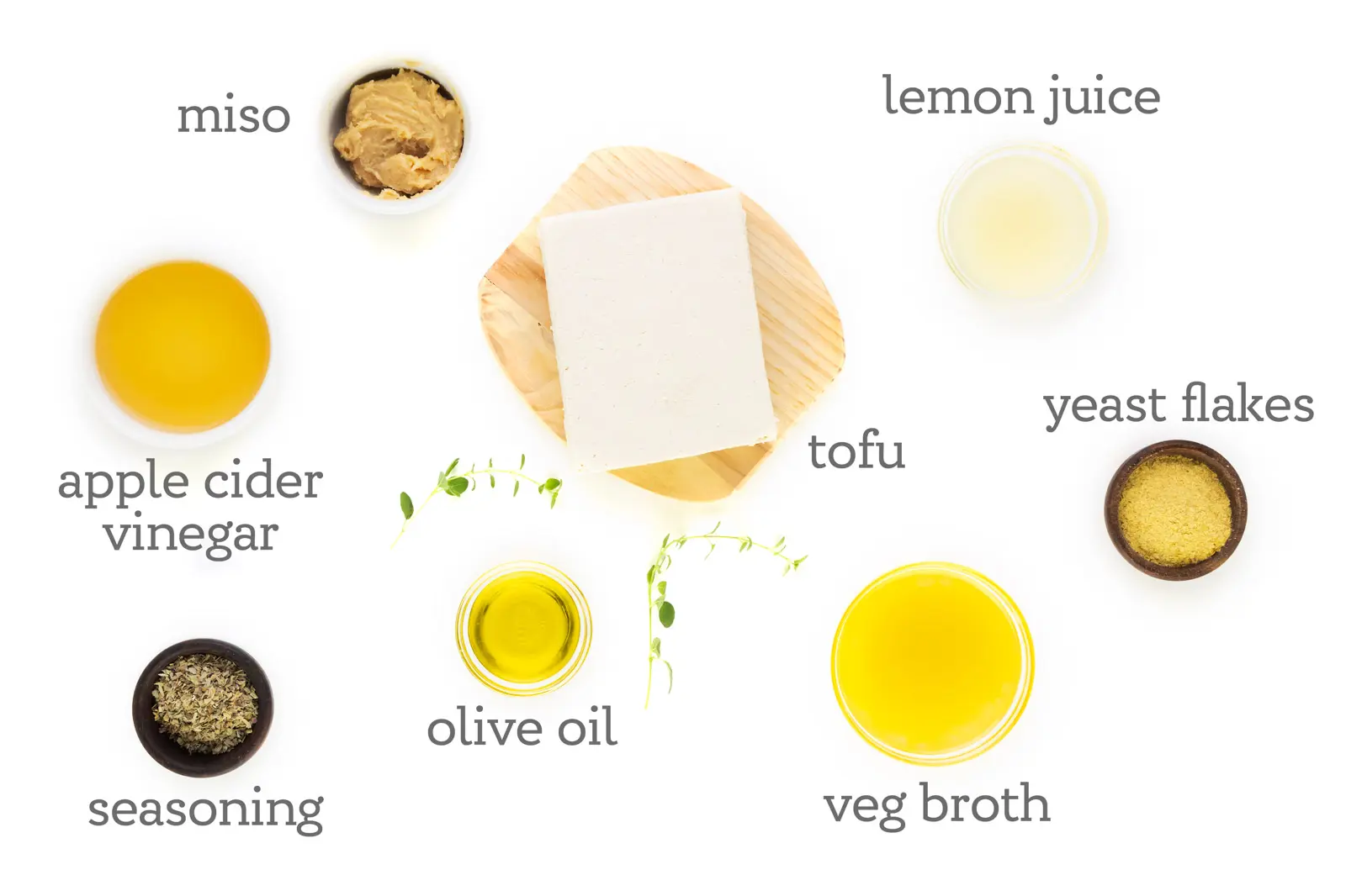 Ingredients for a recipe are grouped on a counter. The labels on them read, "tofu, lemon juice, yeast flakes, veg broth, tofu, olive oil, seasoning, apple cider vinegar, and miso."