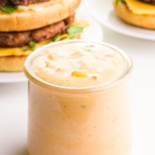 A glass jar of vegan Big Mac special sauce sits in front of two homemade vegan Big Mac burgers.