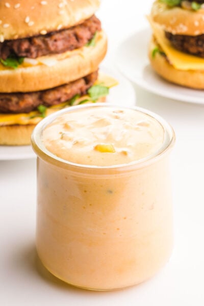 A glass jar of vegan Big Mac special sauce sits in front of two homemade vegan Big Mac burgers.