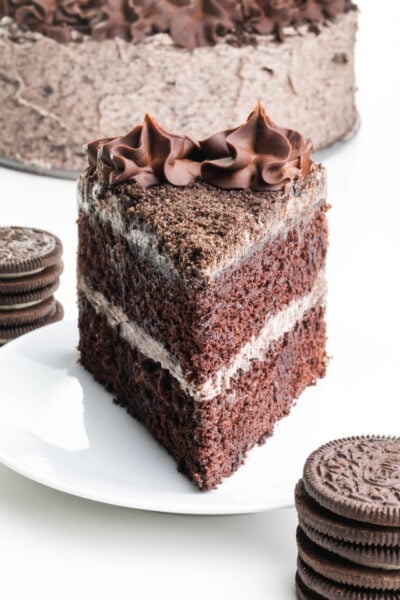 A slice of chocolate cake sits on a plate with chocolate sandwich cookies stacked in front and behind it. The rest of the cake is behind it too.