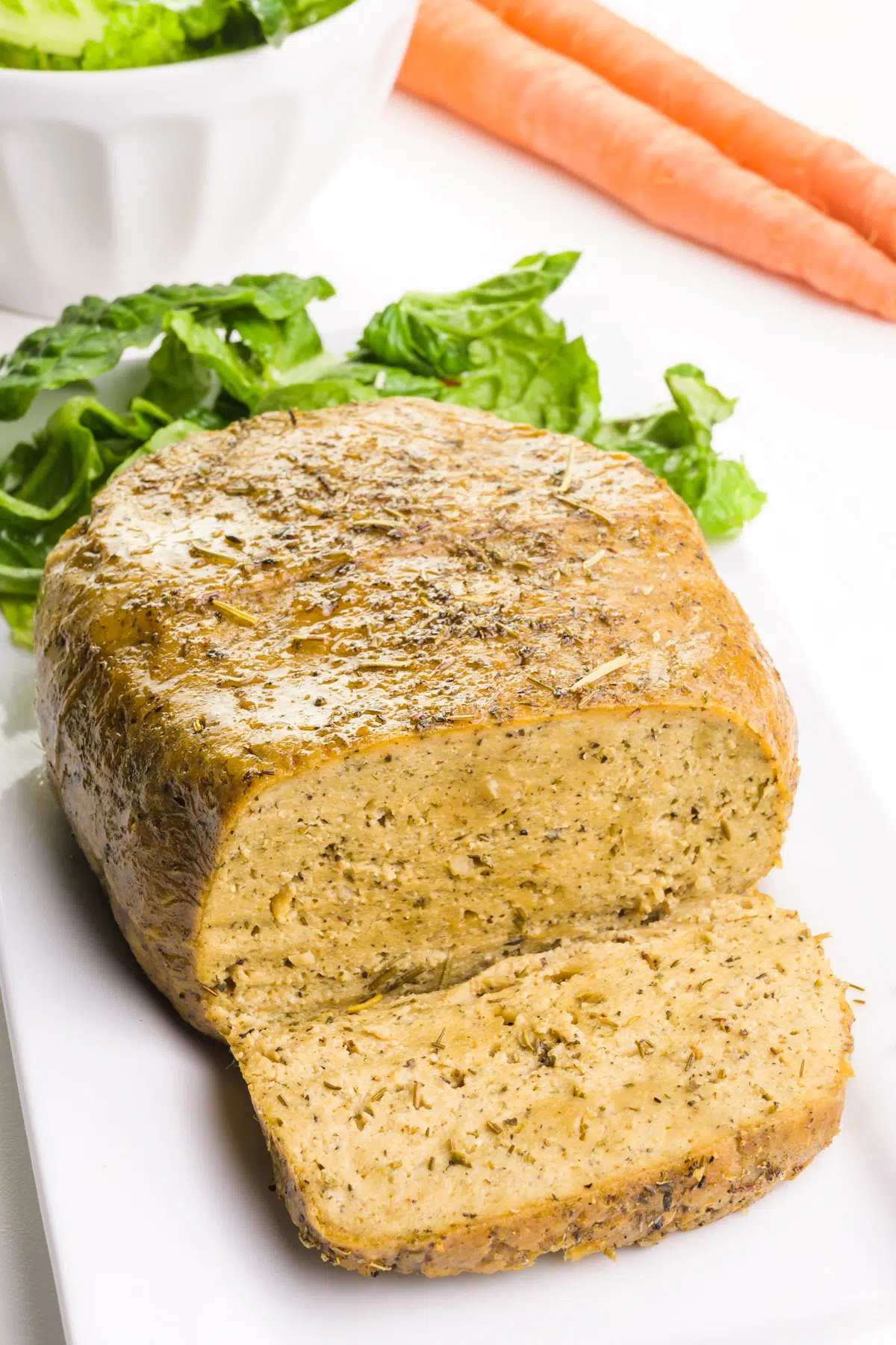 A vegan loaf has a slice cut out and greens in the background along with carrots.