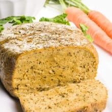 A vegan turkey loaf sits on a plate next to greens and carrots in the background.