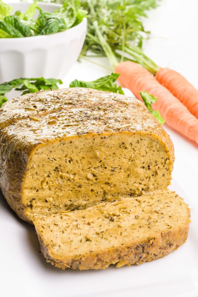 A vegan turkey loaf sits on a plate next to greens and carrots in the background.