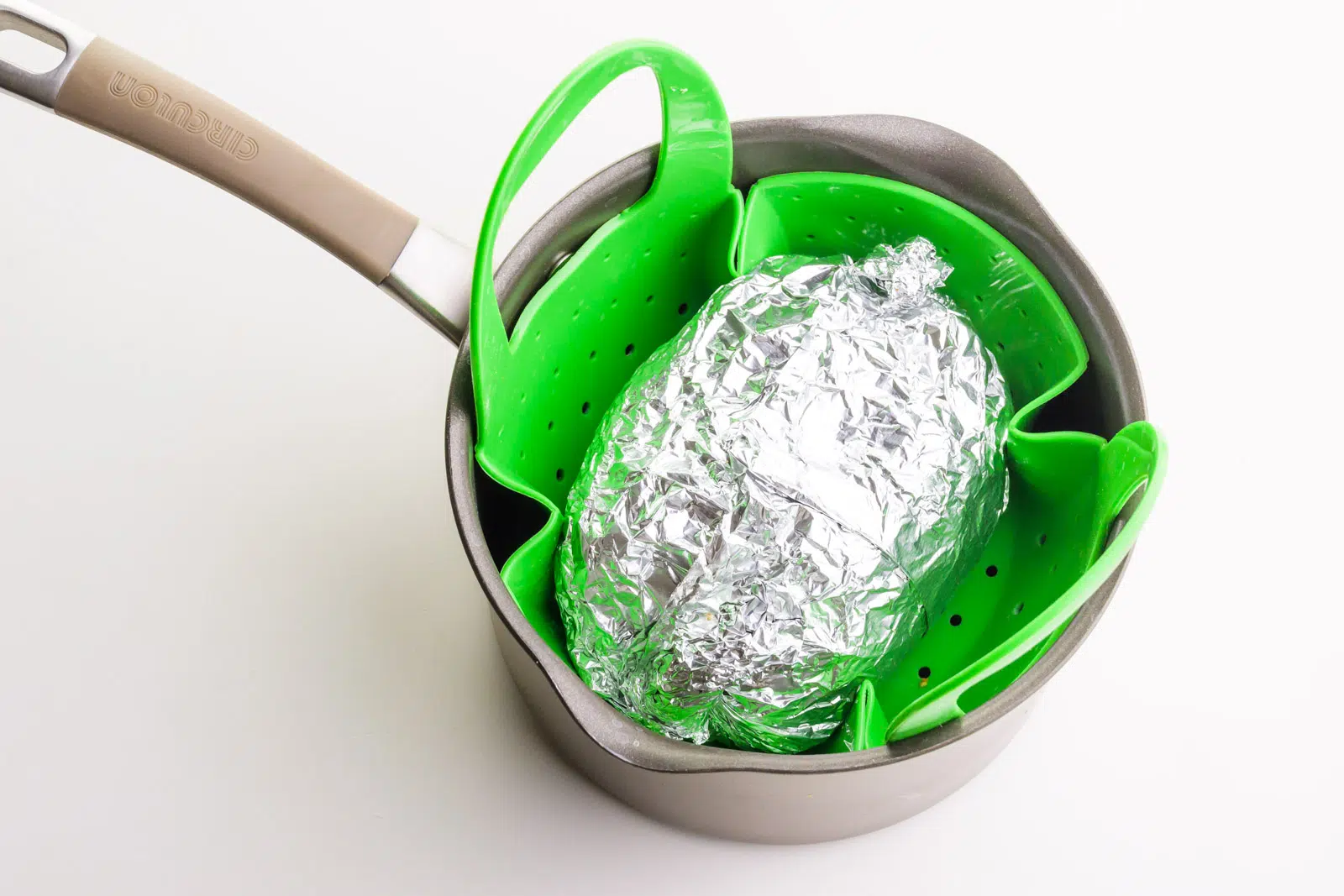 Seitan turkey has been wrapped in foil and is sitting in a steamer basket inside of a saucepan.