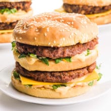 A vegan Big Mac sits on a plate in front of two more sandwiches in the background.