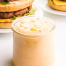 A glass jar of vegan Big Mac special sauce sits in front of two homemade vegan Big Mac burgers.