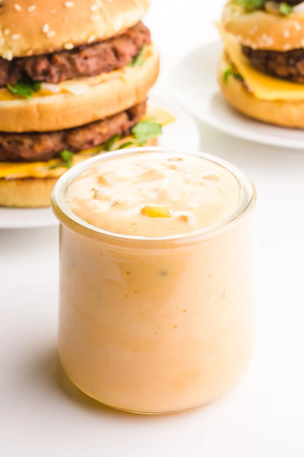 A glass jar of vegan Big Mac special sauce sits in front of two homemade vegan Big Mac burgers.