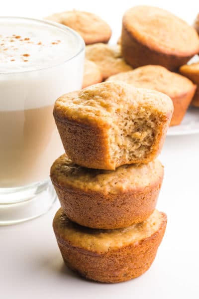 A stack of vegan bran muffins shows a bite taken out of the to one. There's a mug of tea with frothy milk on top and more muffins in the background.