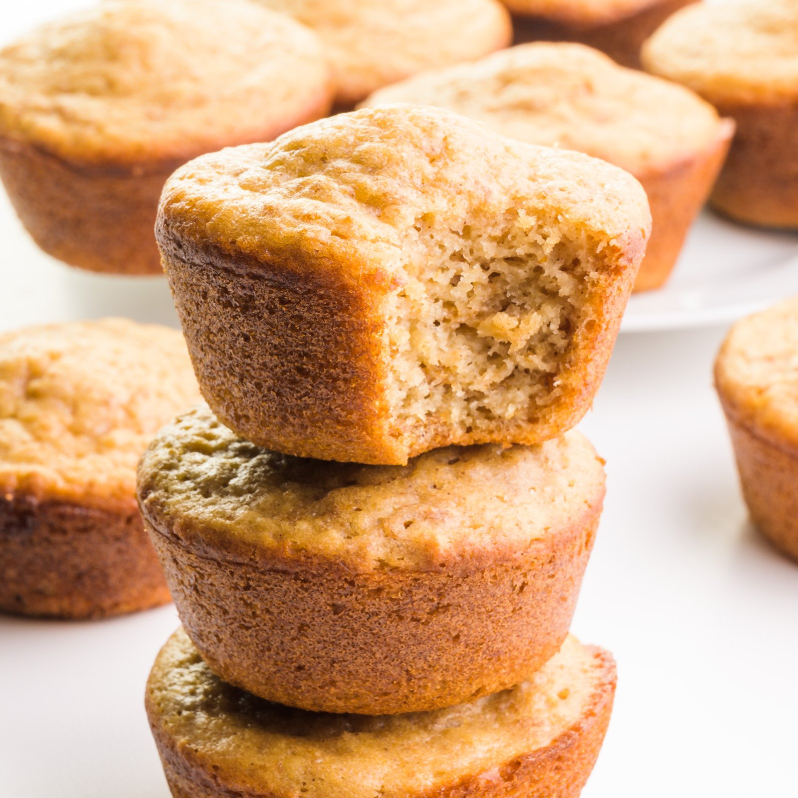 A stack of muffins shows the top one with a bite taken out. There are more muffins in the background.