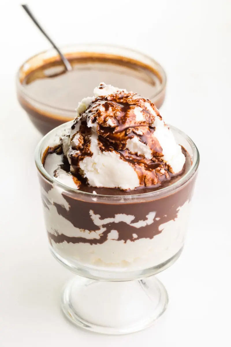 A glass bowl has ice cream with chocolate syrup over the top and the bowl behind it has a spoon in it.