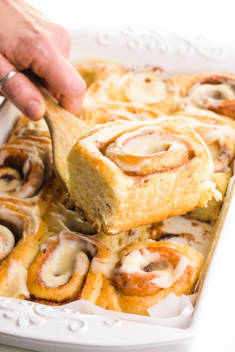A hand holds a spatula with a cinnamon roll on top over a pan with the rest of the rolls.