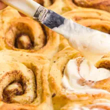 A butter knife is used to drizzle frosting over freshly baked cinnamon rolls.