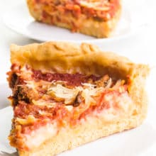 A slice of vegan deep-dish pizza sit on a plate. Another slice is on a plate behind it with the rest of the pizza.