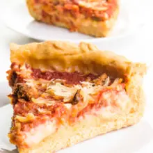 A slice of vegan deep-dish pizza sit on a plate. Another slice is on a plate behind it with the rest of the pizza.
