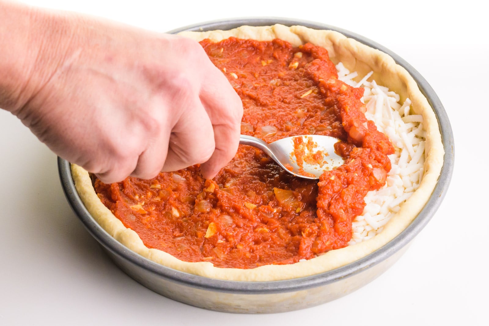 A hand holds a spoon, spreading tomato sauce over a pizza.