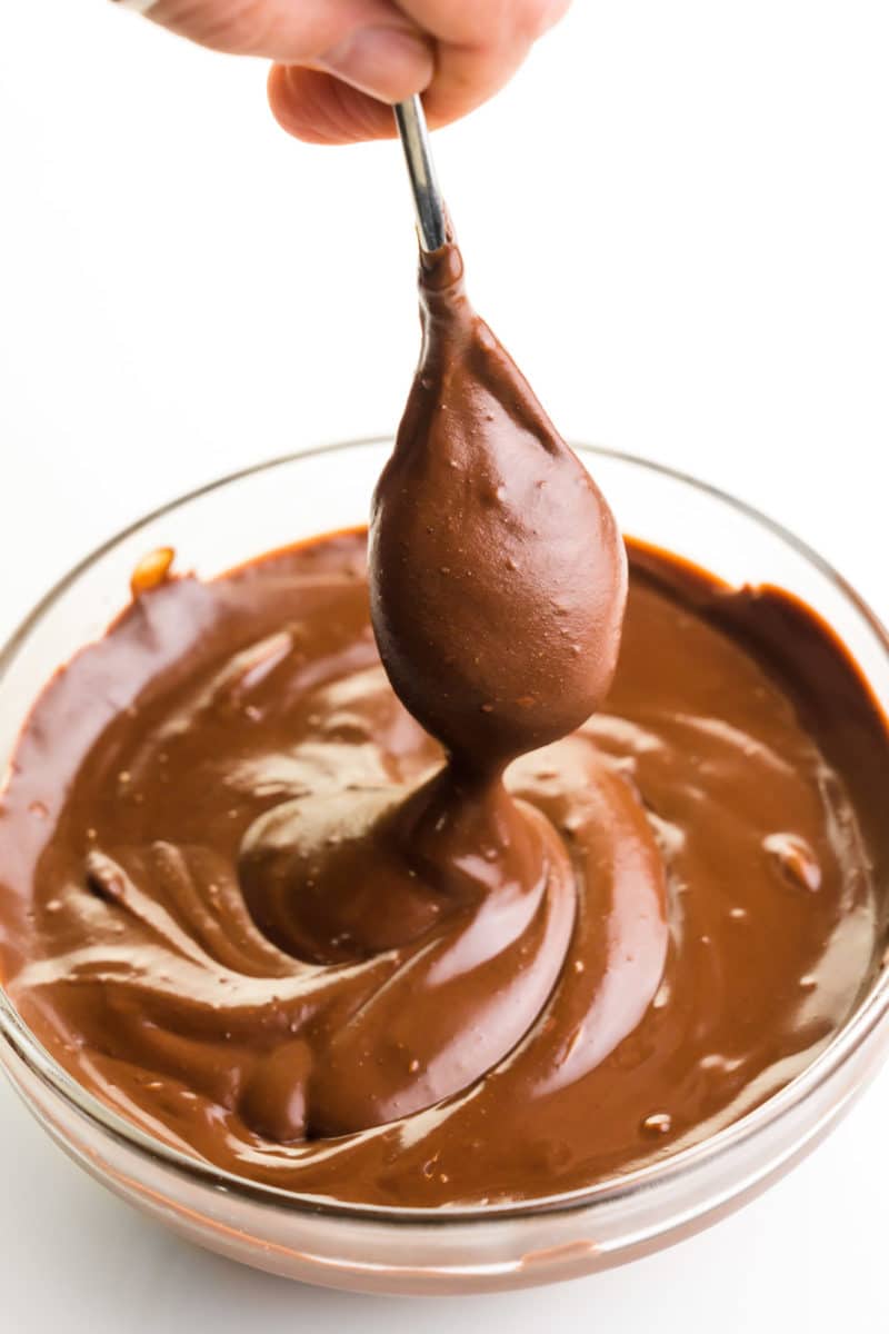 A spoon hovers over a bowl of vegan chocolate ganache. drizzling the chocolate sauce back into the bowl.