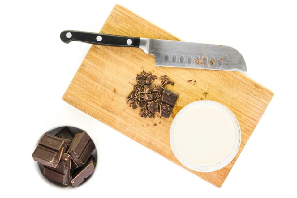 Chocolate chunks are in a white bowl. There are more pieces of chocolate on a cutting board next to a knife and a bowl of coconut milk.