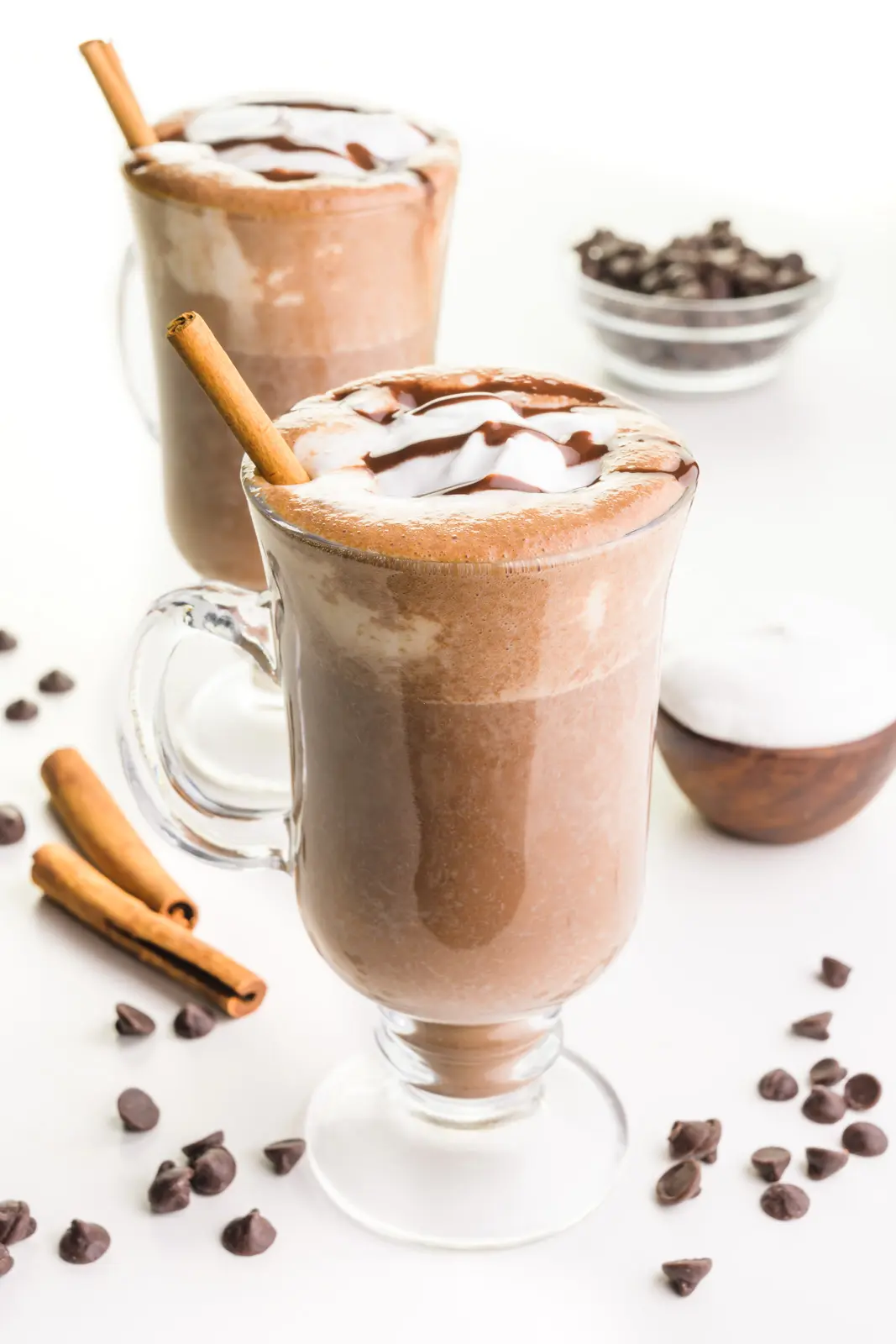 Two glass mugs hold vegan hot chocolate both with cream on top and cinnamon sticks. There are chocolate chips and cinnamon sticks around the mugs and a bowl of whipped cream and chocolate chips.
