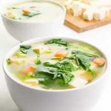 A bowl of vegan miso soup shows pieces of tofu, greens, carrots and more on top. A cutting board in the background has more ingredients, like tofu and green onions.