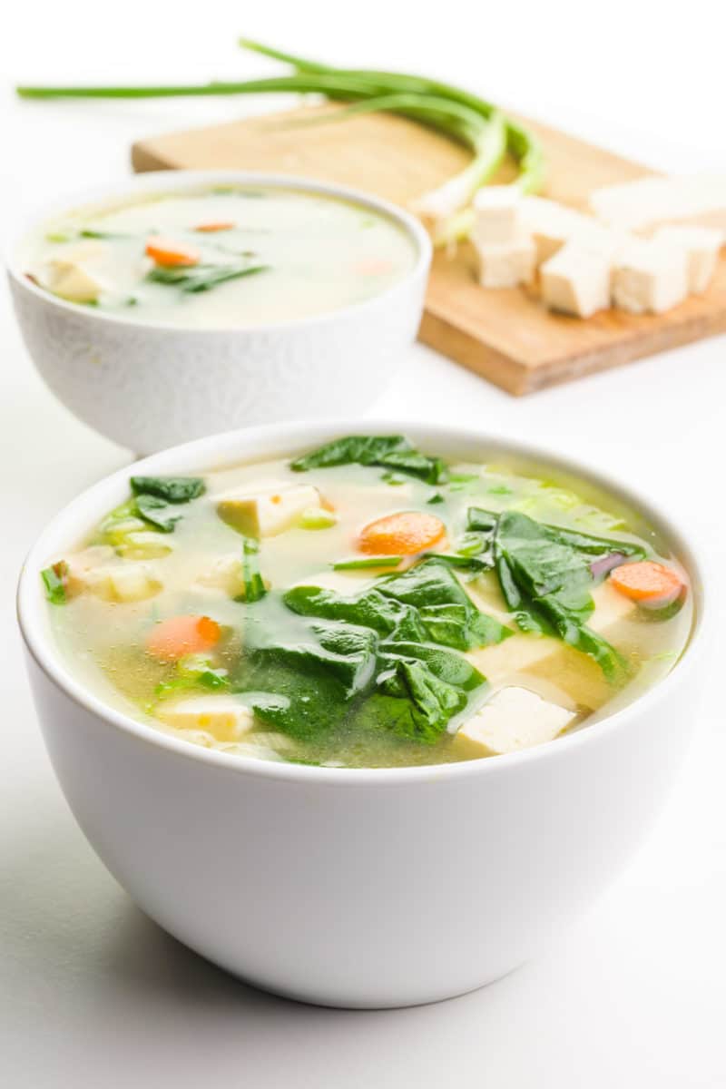 A bowl of vegan miso soup shows pieces of tofu, greens, carrots and more on top. A cutting board in the background has more ingredients, like tofu and green onions.