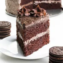 A slice of vegan Oreo cake has stacks of Oreo cookies around it and the rest of the cake behind it.