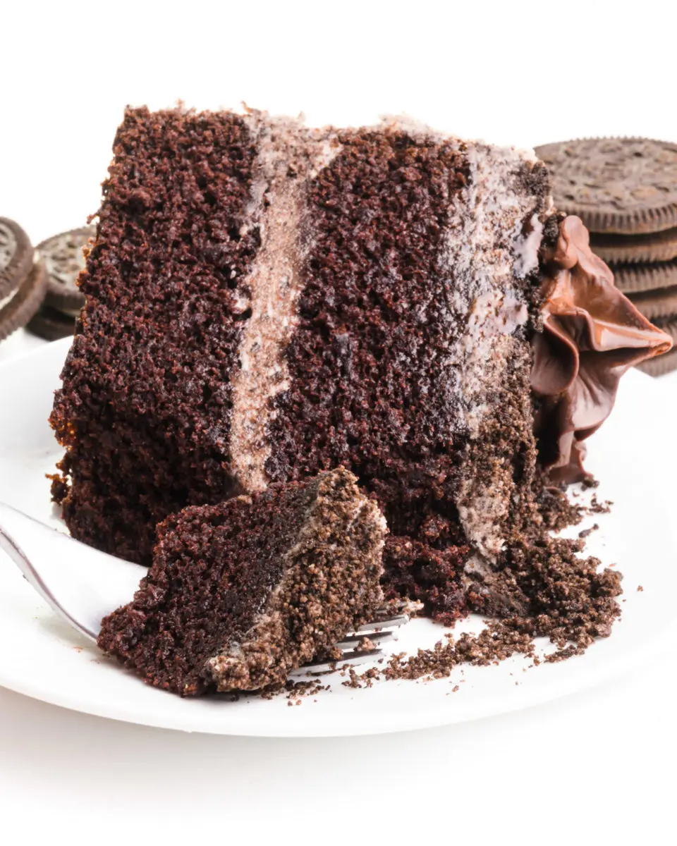 A slice of Oreo cake is on its side and a piece of it is on a fork, showing the frosting. There are cookies stacked in the background.